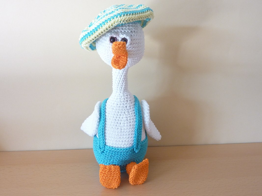 Weißes gehäkeltes Gänse-Amigurumi mit blauer Latzhose, gestreiftem Hut und orangefarbenem Schnabel und Füßen.