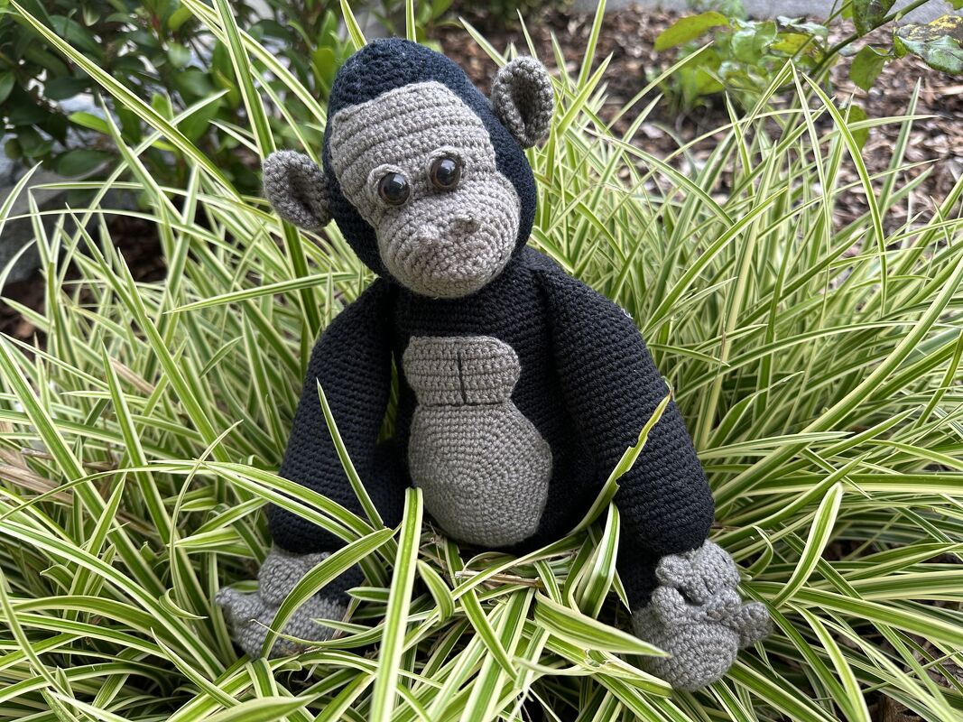 Gehäkelter Amigurumi-Affe in Grau und Dunkelblau sitzt im Ziergras zwischen langen, grün-weiß gestreiften Blättern.