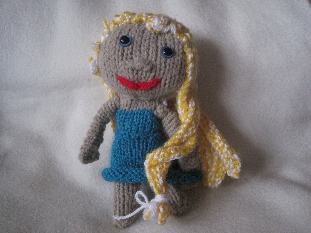 Gestrickte Amigurumi-Puppe mit beigem Körper, blauem Kleid, gelb-weißen Zöpfen, blauen Knopfaugen und rotem Mund