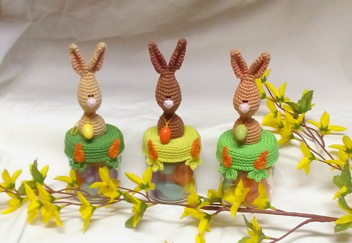 Drei gehäkelte Hasenaufsätze in Braun- und Beigetönen auf Gläsern mit Süßigkeiten, dekoriert mit gelben Forsythienzweigen.