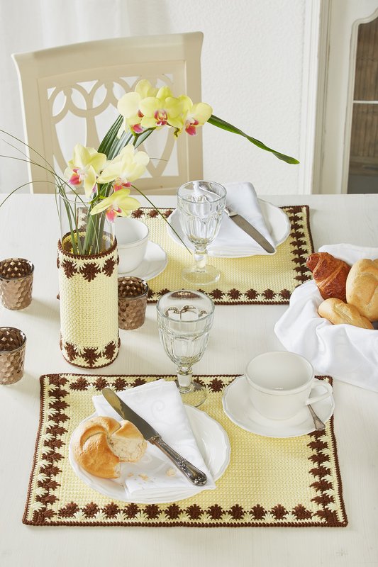 Weißer Esstisch mit zwei gelb-braunen gehäkelten Platzsets, gehäkelter Vasenhülle mit Orchideen, Gläser, Tasse und Brötchen
