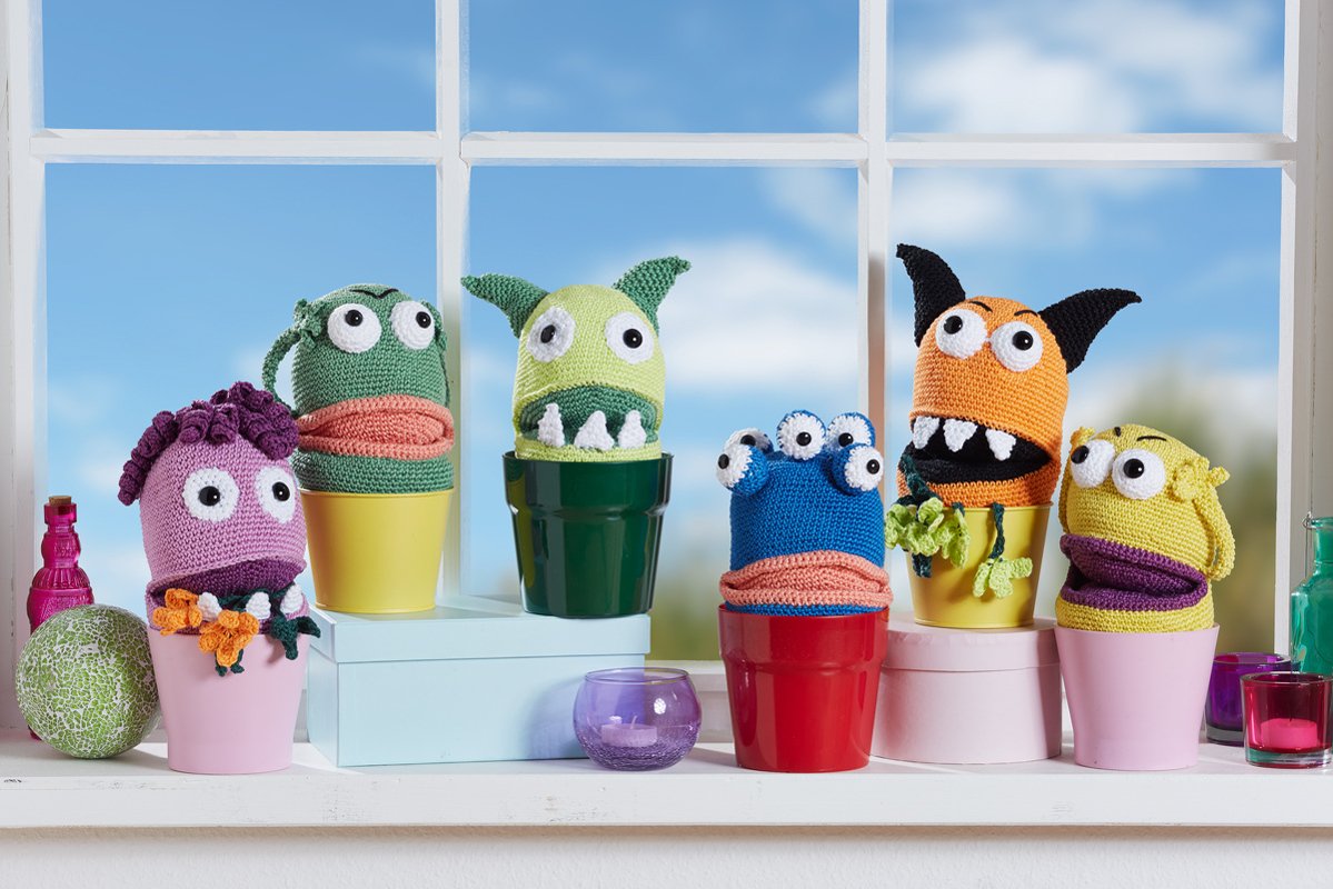 Sechs gehäkelte Amigurumi‑Monster in bunten Blumentöpfen auf einer Fensterbank