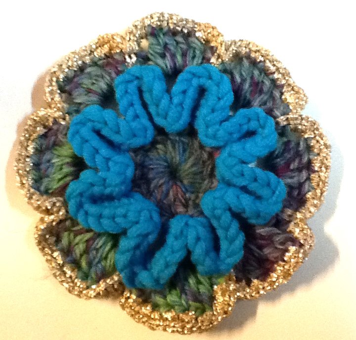 Nahaufnahme einer gehäkelten Blumenapplikation in Blau, Grün und Lila mit goldfarbenem Rand.