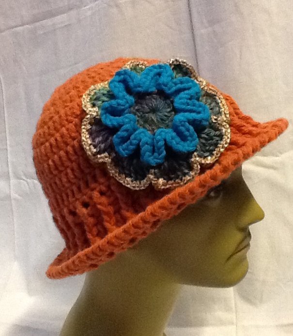 Orangefarbener gehäkelter Hut mit großer blauen und grünen Blütenapplikation auf einem Kopfmodell.