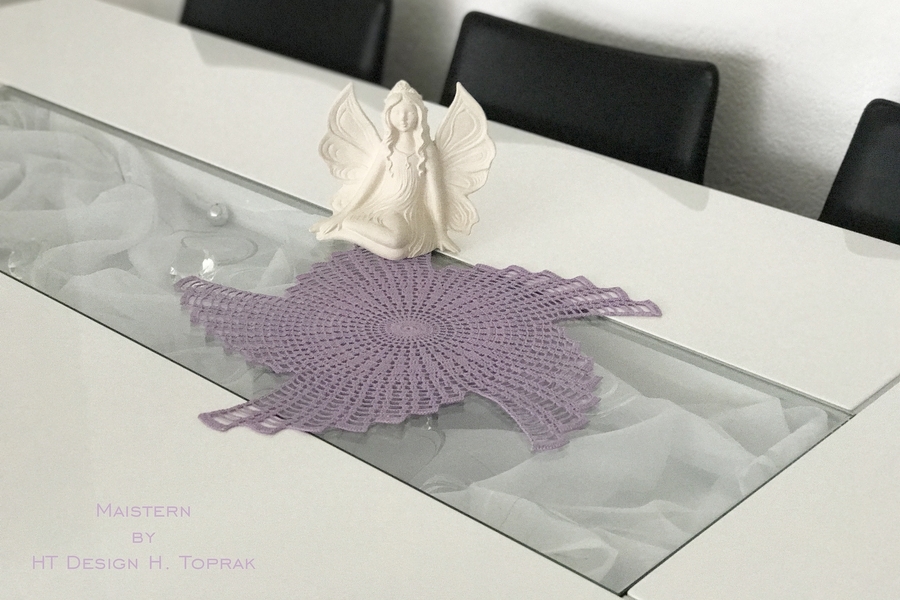Lavendelfarbenes, sternförmig gehäkeltes Deckchen auf einem Glastisch neben einer Engelsfigur.