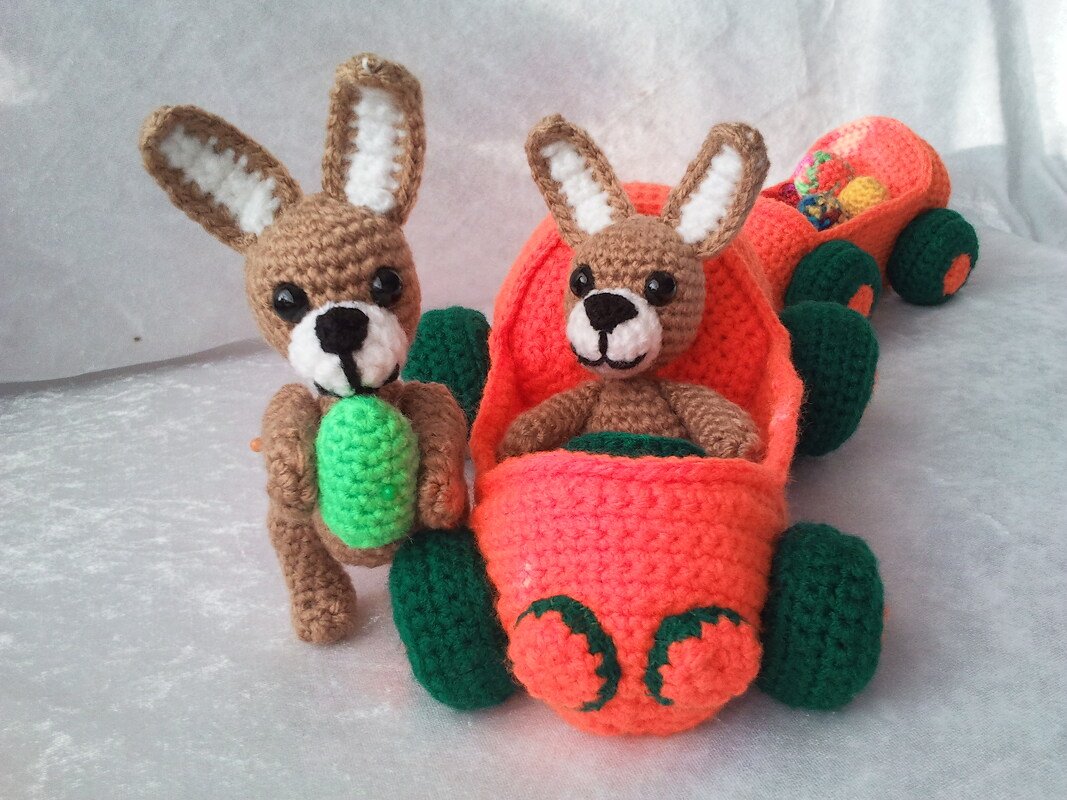 Zwei gehäkelte Amigurumi-Häschen, eines steht mit grünem Ei, das andere sitzt im orangefarbenen Karottenauto mit Anhänger voller Eier.