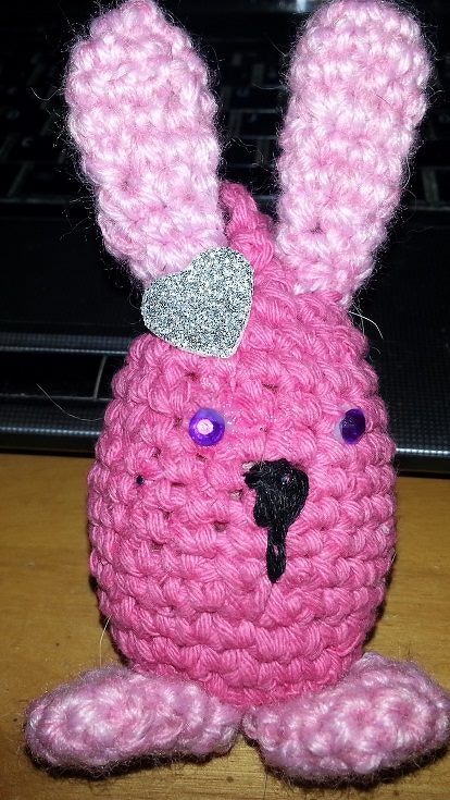 kleines rosa Amigurumi-Häschen mit sichtbaren Maschen, violetten Knopfaugen und glitzerndem Herz am Ohr