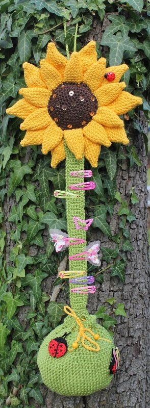 Gehäkelter Sonnenblumen-Organizer mit gelben Blütenblättern, braunem Pailletten-Kern, Marienkäfer-Applikationen, grünem Stiel mit bunten Haarspangen und Beutel, vor Efeu an einem Baumstamm.