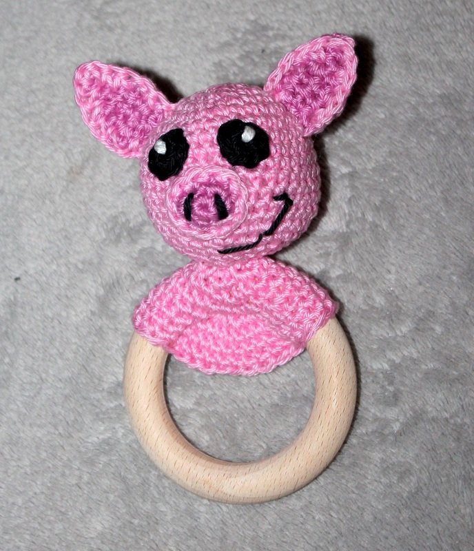 Rosa gehäkeltes Schweinchen als Greifling mit hellem Holzring auf grauem Stoff