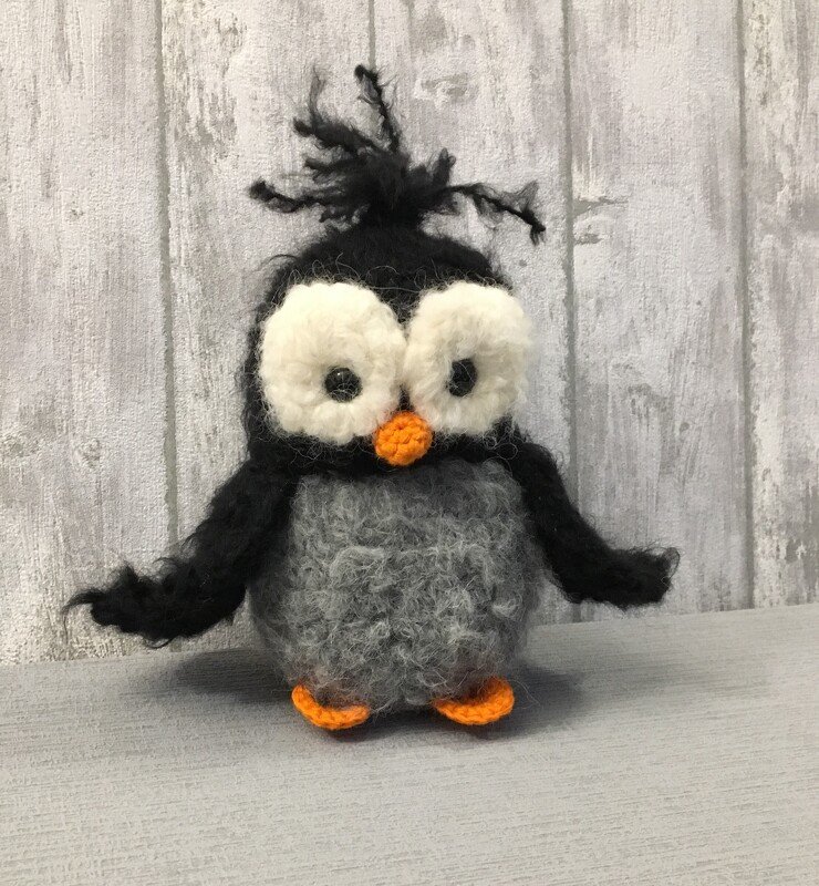 Gehäkelter Amigurumi-Pinguin in Schwarz und Grau mit großen weißen Augen und orangefarbenem Schnabel und Füßen.