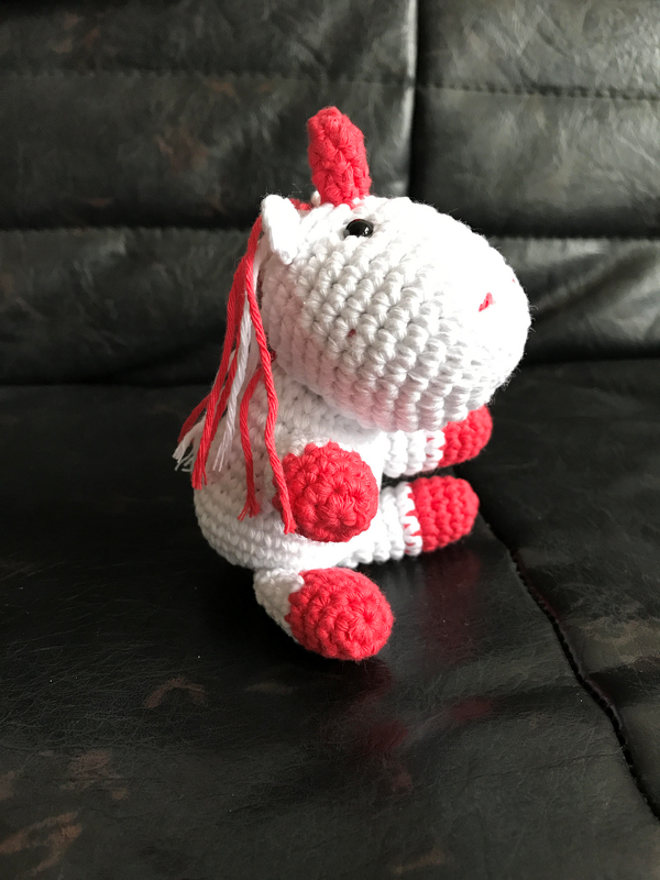 Gehäkeltes Einhorn-Amigurumi in Weiß mit roten Hufen und Horn, Seitenansicht auf dunklem Untergrund