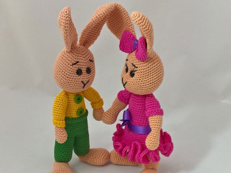 Zwei gehäkelte Amigurumi-Hasen halten Händchen; gelbes Oberteil mit grüner Hose und pinkes Kleid mit Schleife
