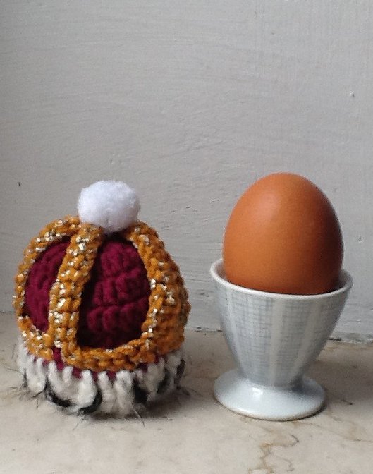 Gehäkelter Eierwärmer in Kronenform (Burgunder, Gold, Weiß) mit Pompon neben braunem Ei im Eierbecher.