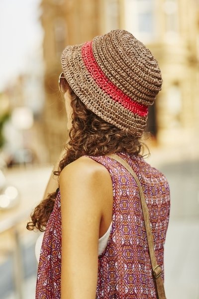 Frau von hinten mit beigem gehäkelten Hut mit roten Streifen, lockigem Haar und gemusterter Bluse.