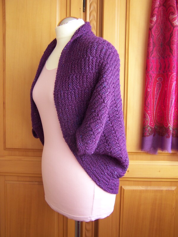 Mannequin mit lilafarbenem, gestricktem Seelenwärmer und kurzen Ärmeln vor Holztür