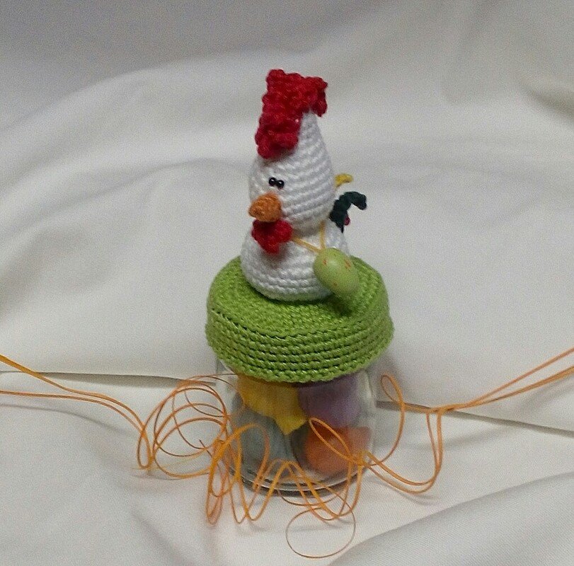 Glas mit grünem Häkeldeckel, darauf eine weiße gehäkelte Henne mit rotem Kamm; Glas gefüllt mit pastellfarbenen Eiern und oranger Bastdeko