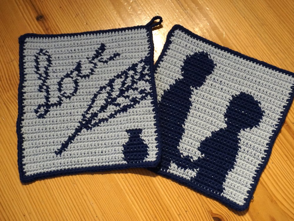 Zwei gehäkelte, quadratische Topflappen in Hell- und Dunkelblau: einer mit dem Schriftzug „love“ und floralem Motiv, der andere mit Pärchen-Silhouette.