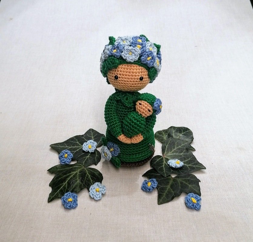 Gehäkelte Amigurumi-Puppe im grünen Outfit mit blau-weißem Blumenkranz, die ein kleines Baby im Arm hält, umgeben von Efeublättern und Häkelblüten.