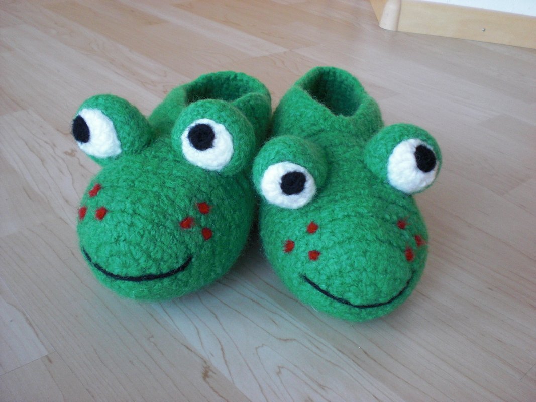 Paar grüne, gehäkelte Filz-Hausschuhe in Froschform mit großen Augen und lächelndem Mund auf hellem Holzboden.
