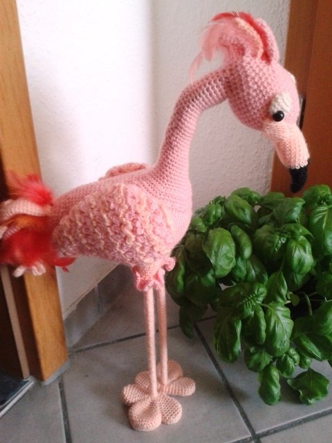 Gehäkelter rosa Flamingo-Amigurumi mit langen Beinen und schwarzer Schnabelspitze neben Topfpflanze