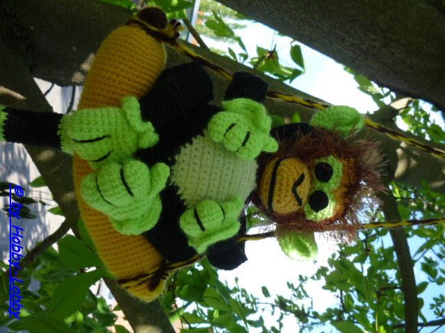 Gehäkelter Amigurumi-Affe in Schwarz und Hellgrün mit brauner Mähne, kopfüber an einem Ast hängend und eine gelbe Banane haltend.
