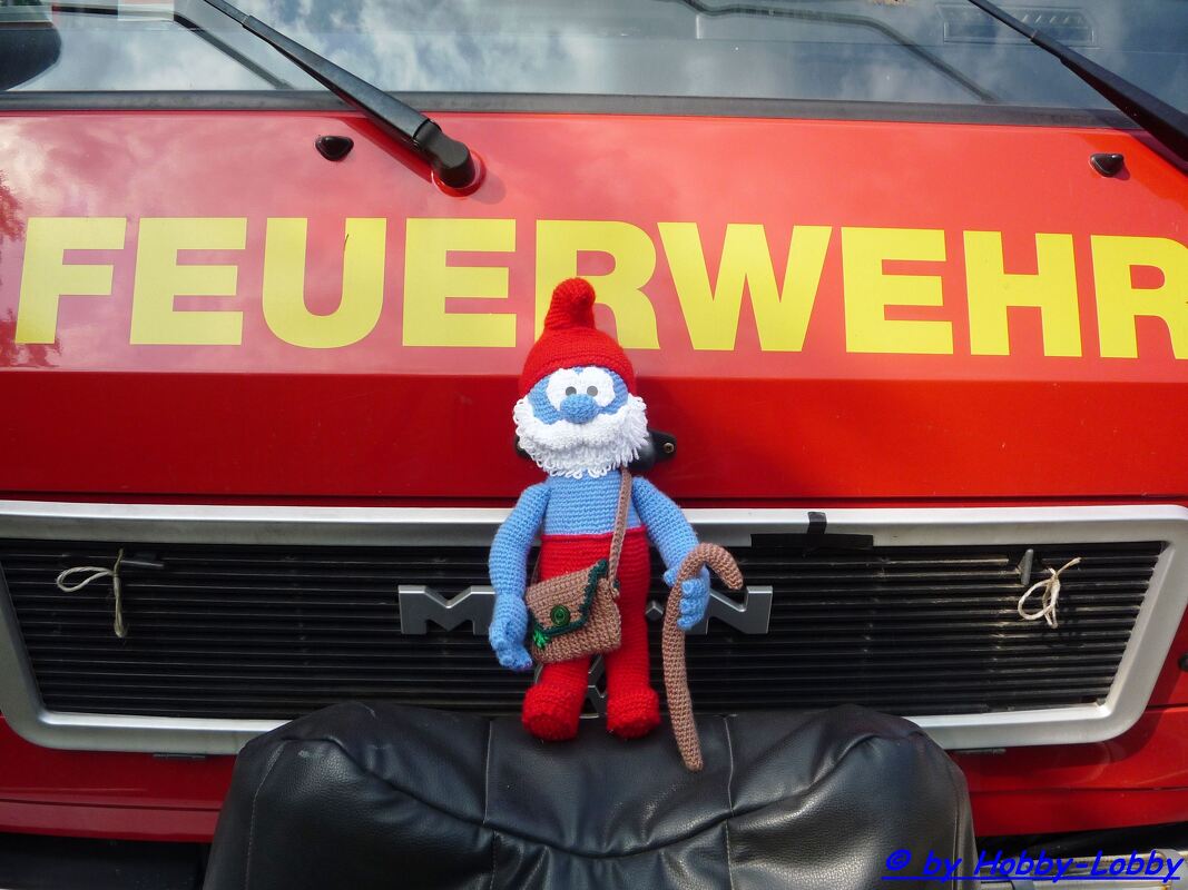 Gehäkelte Papa Schlumpf-Figur mit rotem Hut, weißem Bart und Gehstock vor einem roten Feuerwehrwagen mit Schriftzug FEUERWEHR.
