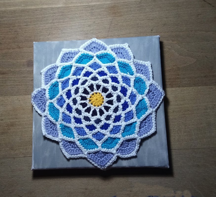 Rundes handgehäkeltes Mandala in Blau-, Türkis- und Violetttönen mit gelber Mitte auf grauer Leinwand