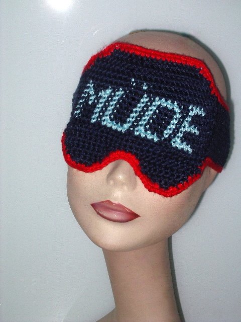 Gehäkelte dunkelblaue Schlafmaske mit rotem Rand und hellblauem Schriftzug 'MÜDE' auf einer Schaufensterpuppe.