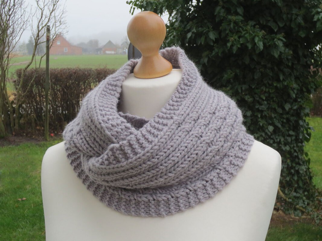 Hellgrauer gestrickter Loop-Schal mit Rippen- und Strukturmuster auf einer weißen Schaufensterpuppe vor unscharfem Außenhintergrund.