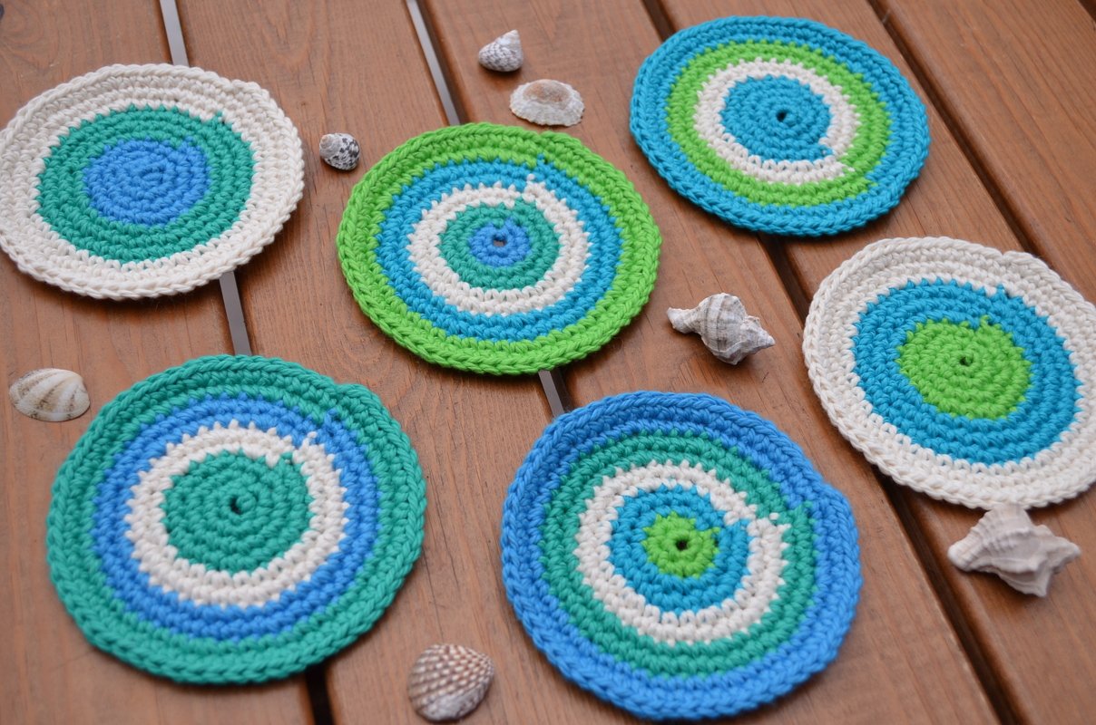 Sechs runde, handgehäkelte Untersetzer in Blau, Grün und Weiß auf Holz, umgeben von kleinen Muscheln.