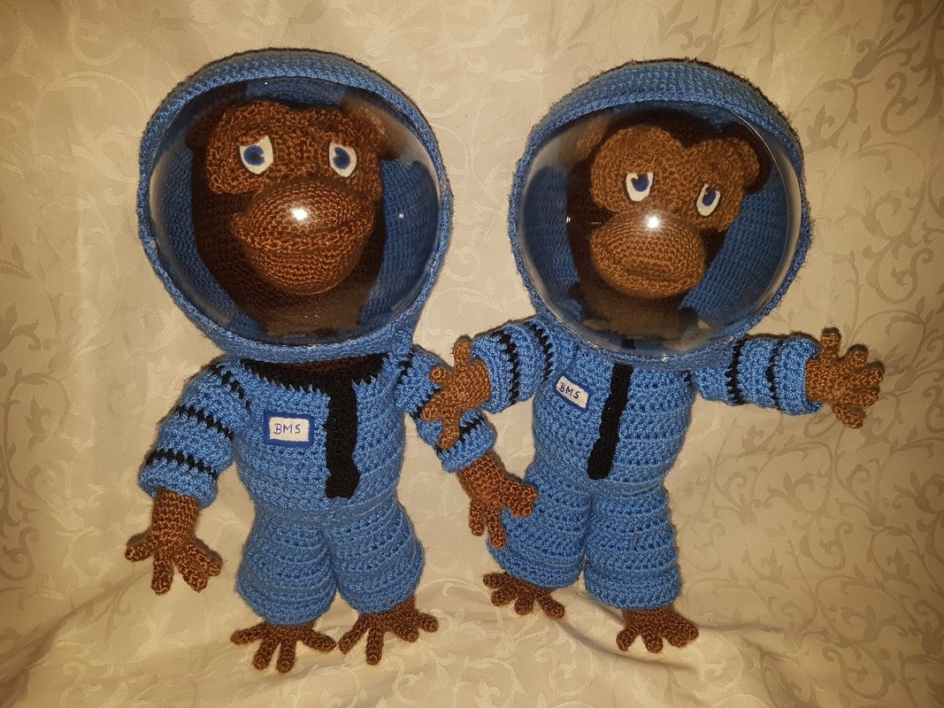 Zwei gehäkelte Affen in blauen Astronautenanzügen mit klaren Helmen, Händchen haltend, auf hellem Hintergrund