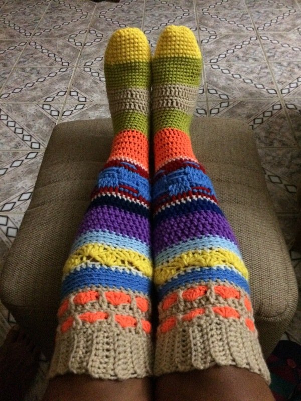 Feet and calves wearing long, striped crocheted leg warmers in yellow, green, orange, blue, purple and beige, resting on a brown cushion with tiled floor visible.