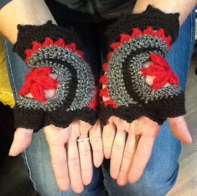 Paar gehäkelter fingerloser Armstulpen in Schwarz, Grau und Rot, getragen an geöffneten Händen