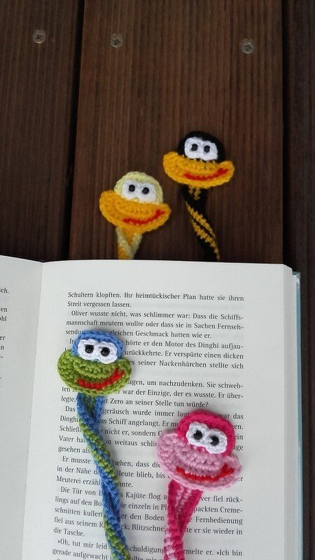 Fünf gehäkelte Tier-Lesezeichen, zwei auf Holz, drei in einem offenen Buch, verschiedene Farben