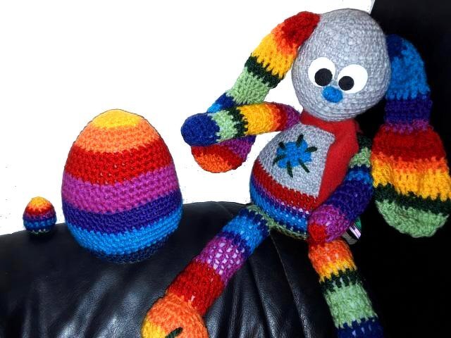 Gehäkelter Hase in Regenbogenstreifen neben zwei gestreiften Häkel-Eiern