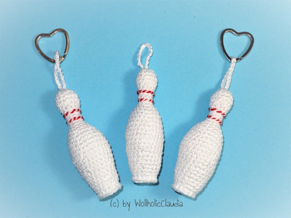 Drei gehäkelte weiße Kegel‑Schlüsselanhänger mit rot‑weißem Zierfaden und zwei herzförmigen Ringen auf hellblauem Hintergrund.