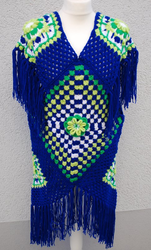 Blauer gehäkelter Poncho aus Granny-Squares mit grünen und weißen Akzenten, V‑Ausschnitt, Fransen und zentraler Häkelblume auf Schaufensterpuppe
