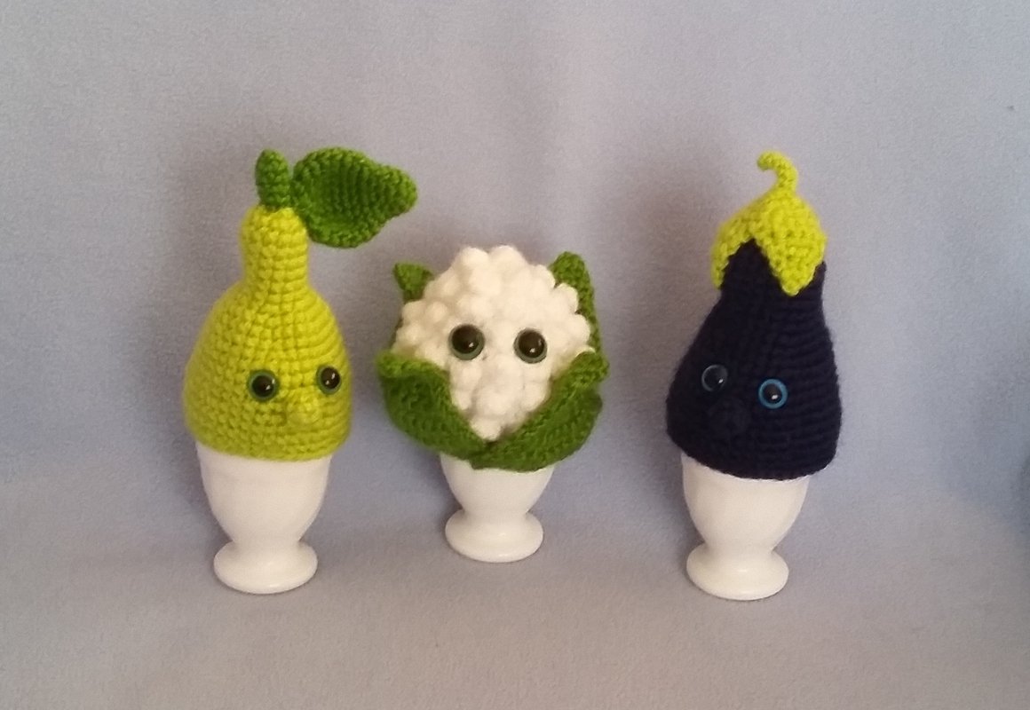 Drei gehäkelte Eierwärmer in Birnen-, Blumenkohl- und Auberginenform auf weißen Eierbechern