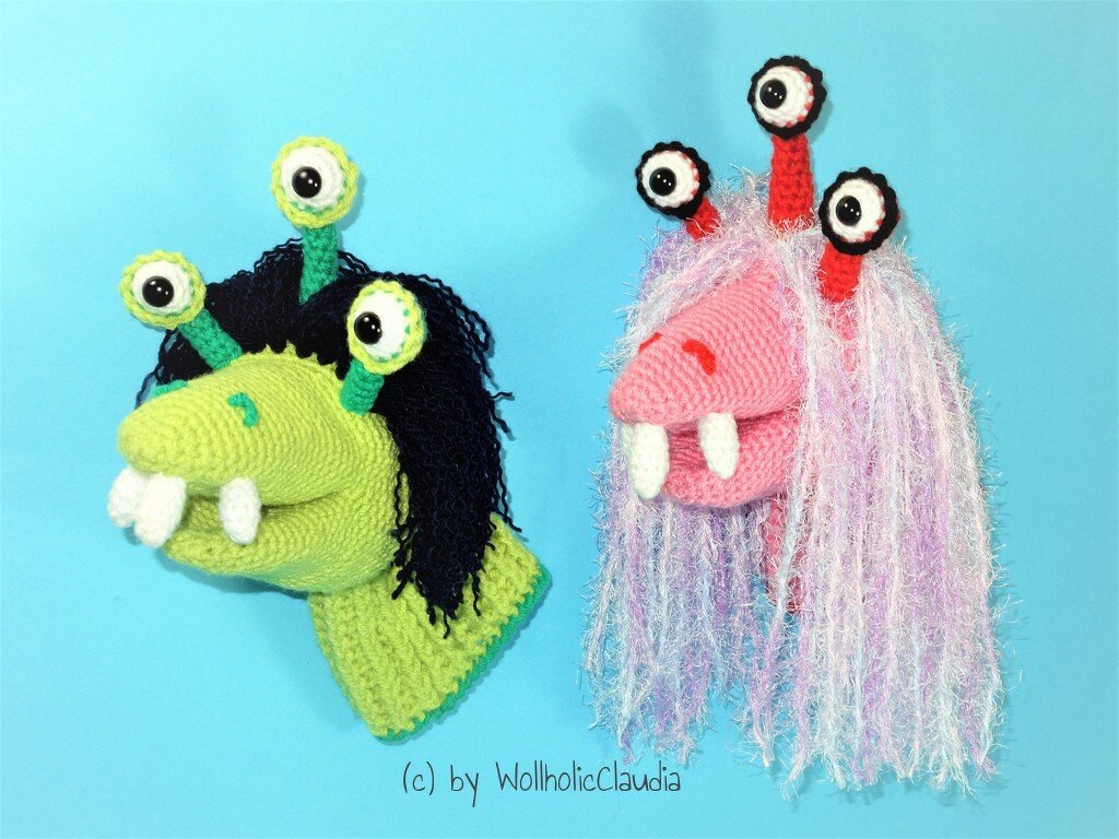 Zwei gehäkelte Amigurumi-Handpuppen: links grün mit schwarzem Haar und drei Augenstielen, rechts rosa mit flauschigem pastellfarbenem Haar und drei Augenstielen