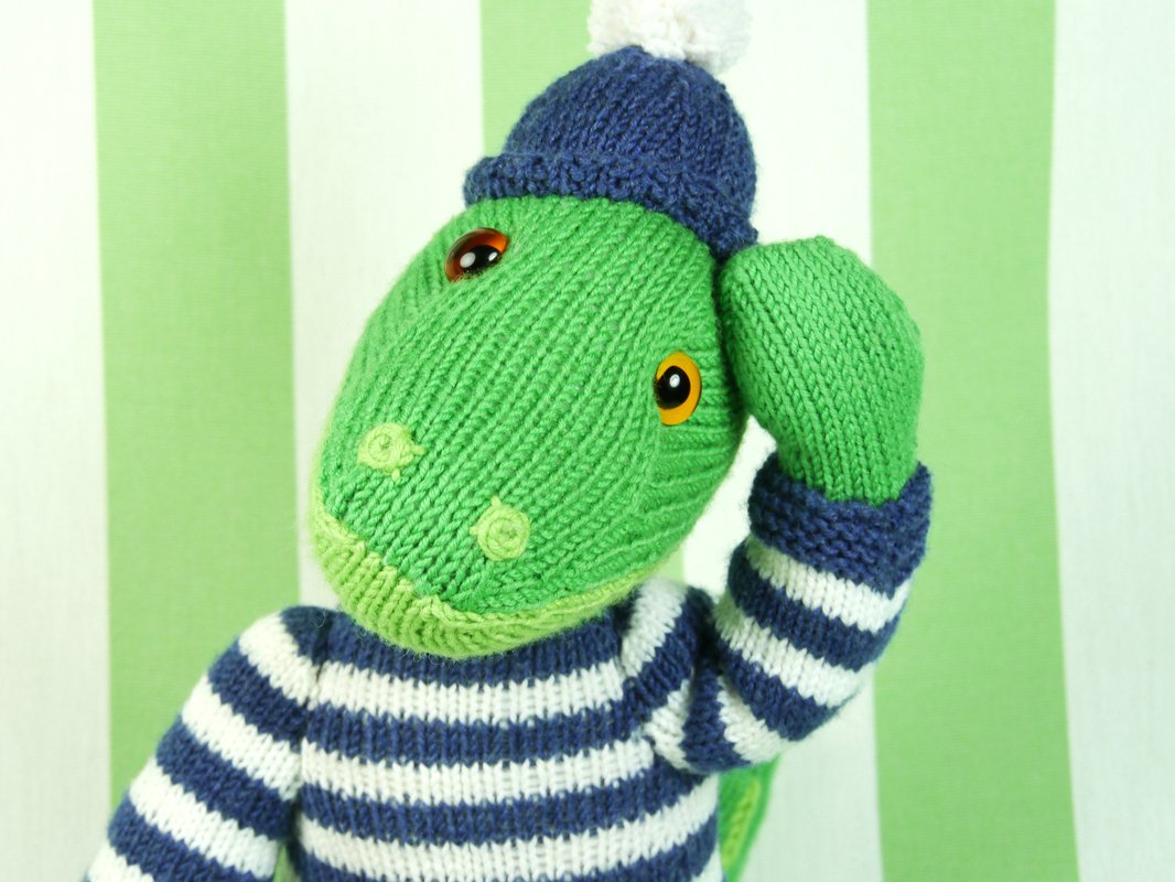 Green knitted crocodile plush wearing a navy-and-white striped sweater and navy beanie with pompom