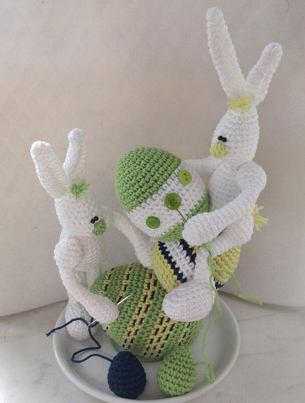 Zwei weiße gehäkelte Amigurumi-Hasen halten ein teilweise zusammengesetztes, mehrfarbiges Osterei mit Nadel, Garn und Knopfdetails