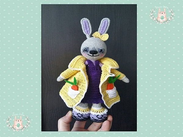 Gehäkelter Amigurumi-Hase mit gelbem Mantel, Karotten in den Taschen und Schleife am Ohr, von Hand gehalten