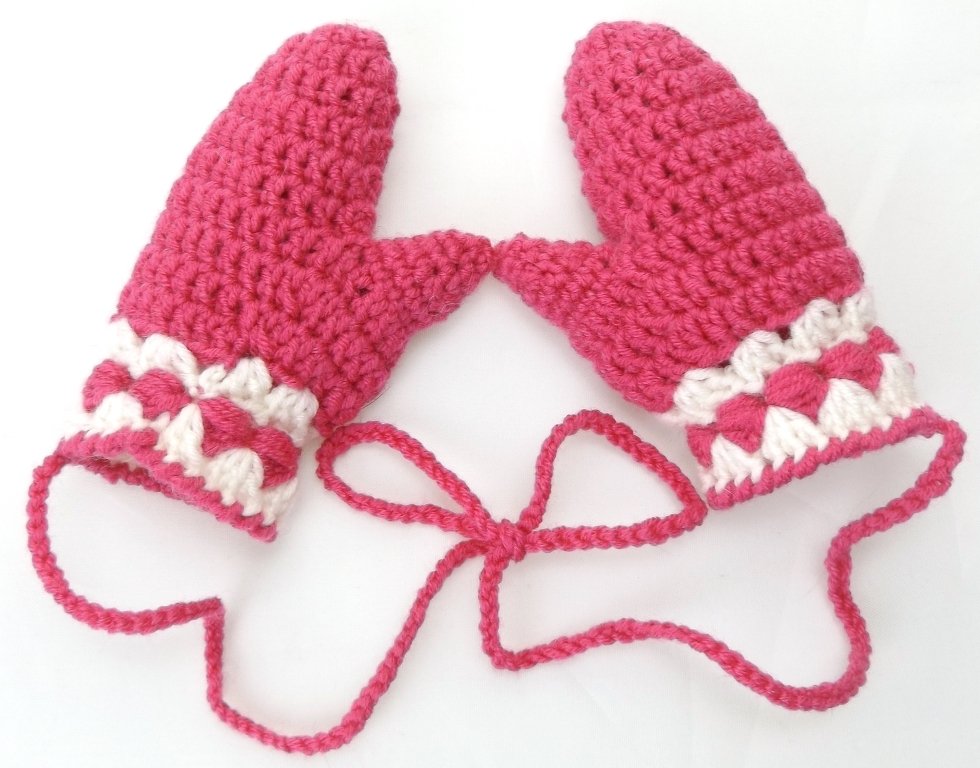 Zwei pinke gehäkelte Babyfäustlinge mit weiß-pinker Zierborte und verbindender Kordel auf hellem Hintergrund.