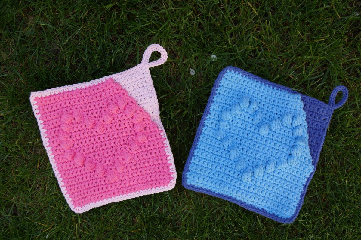 Zwei quadratische gehäkelte Topflappen in Rosa und Hellblau mit Herznoppen auf Gras