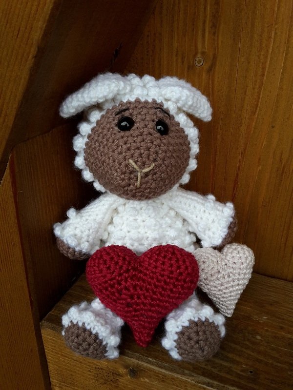 Gehäkeltes Amigurumi‑Schäfchen in Weiß und Braun, hält rotes und beiges Herz