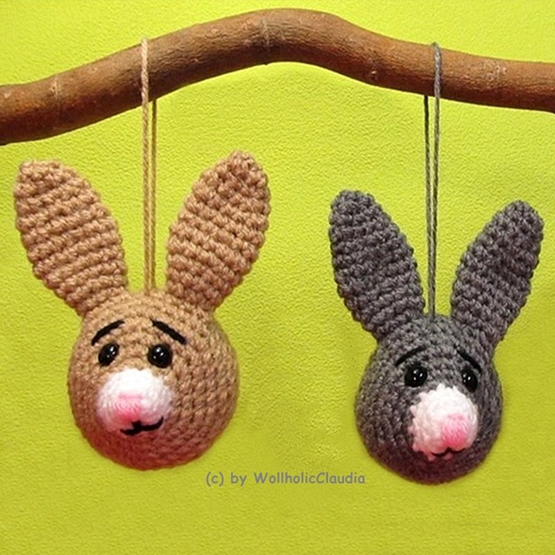 Zwei gehäkelte Hasenkopf-Anhänger, einer braun, einer grau, mit Schlaufe an einem Ast vor grünem Hintergrund