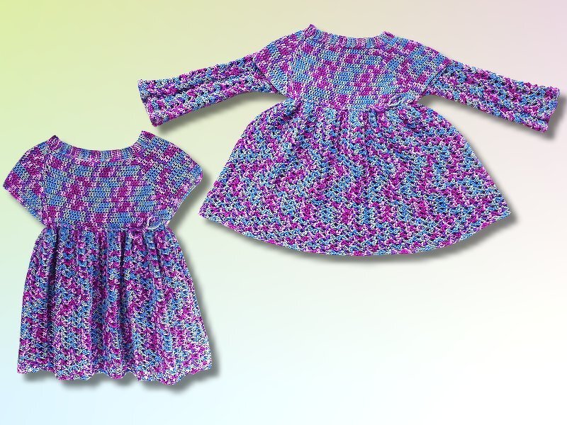 Zwei gehäkelte Kinderkleider in Lila und Blau, eins kurzärmlig, eins langärmlig, nebeneinander gelegt.