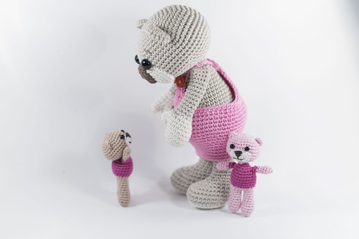 Drei gehäkelte Amigurumi-Figuren: großer hellgrauer Bär mit rosa Latzhose und zwei kleinere Figuren auf weißem Hintergrund