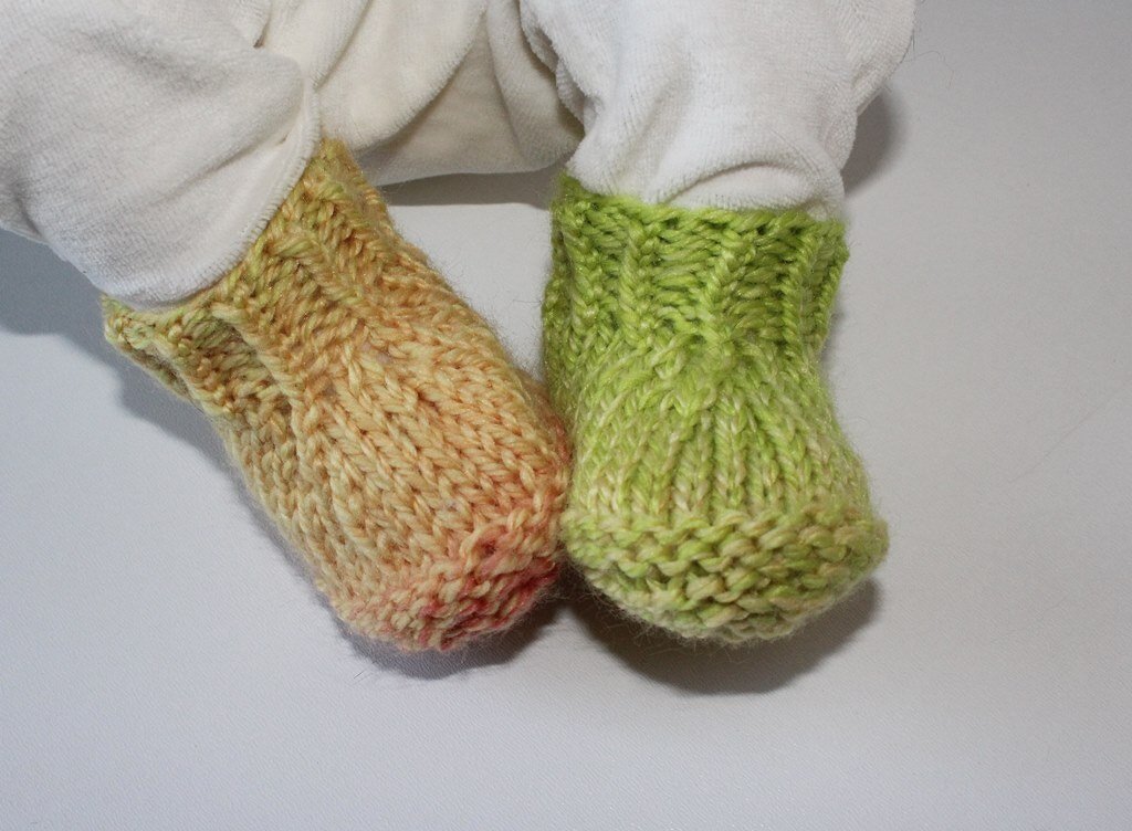 Zwei gestrickte Babyschuhe, einer orange-meliert und einer grün-meliert, an den Füßen eines Babys.