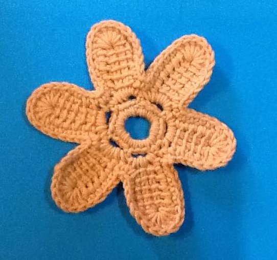 Beige gehäkelte Blumen-Applikation mit sechs Blütenblättern und offenem Zentrum auf blauem Hintergrund